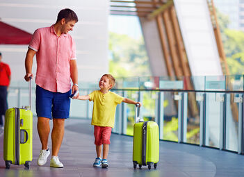 Ein Vater geht mit seinem Kind an der Hand am Flughafen. Der Vater und sein Kind haben einen Koffer in der Hand.