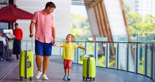 Ein Vater geht mit seinem Kind an der Hand am Flughafen. Der Vater und sein Kind haben einen Koffer in der Hand.