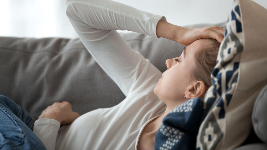 Eine junge Frau liegt erschöpft auf dem Sofa und hält ihre Hand an den Kopf.