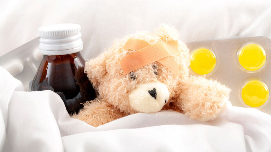 Ein Teddybär liegt zugedeckt im Bett und hat ein Pflaster am Kopf kleben. Neben dem Stofftier liegen Lutschtabletten und ein Hustensaft.