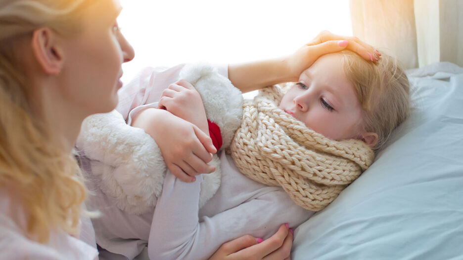 Ein Kind liegt mit einem Schal und einem Teddy-Bären im Bett. Ihre Mutter kümmert sich um das Kind.