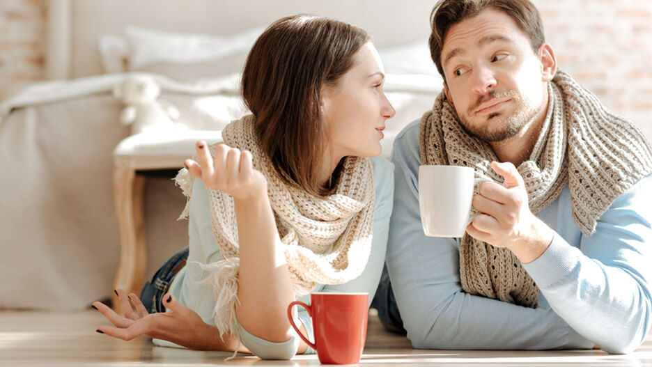 Eine junge Frau und ein Mann liegen auf dem Boden und trinken Tee. Im Hintergrund ist eine Bank und ein Bett zu sehen.