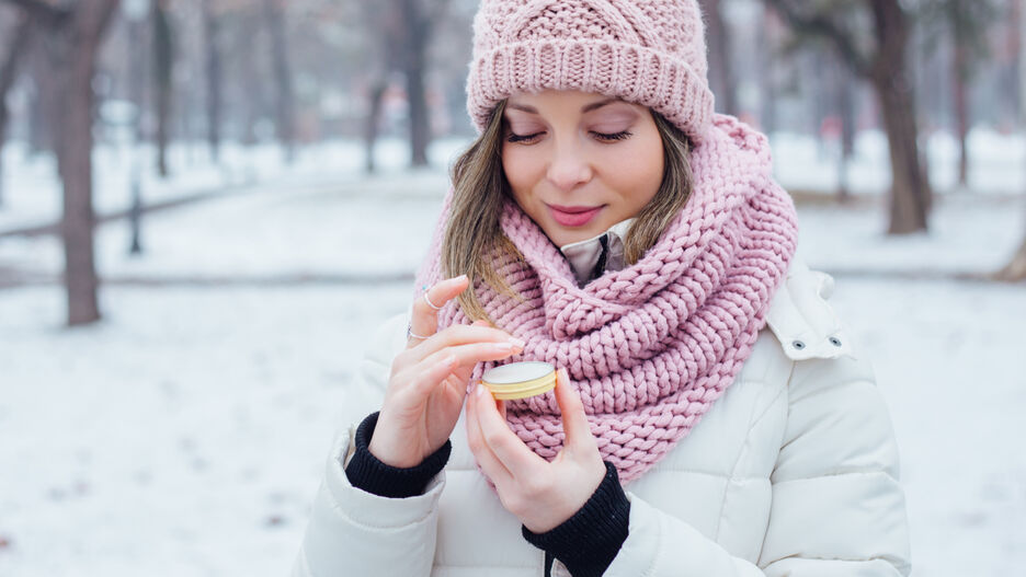 Eine junge Frau steht in Winterkleidung in einer verschneiten Landschaft und trägt Lippenbalsam auf ihre Lippen auf. 