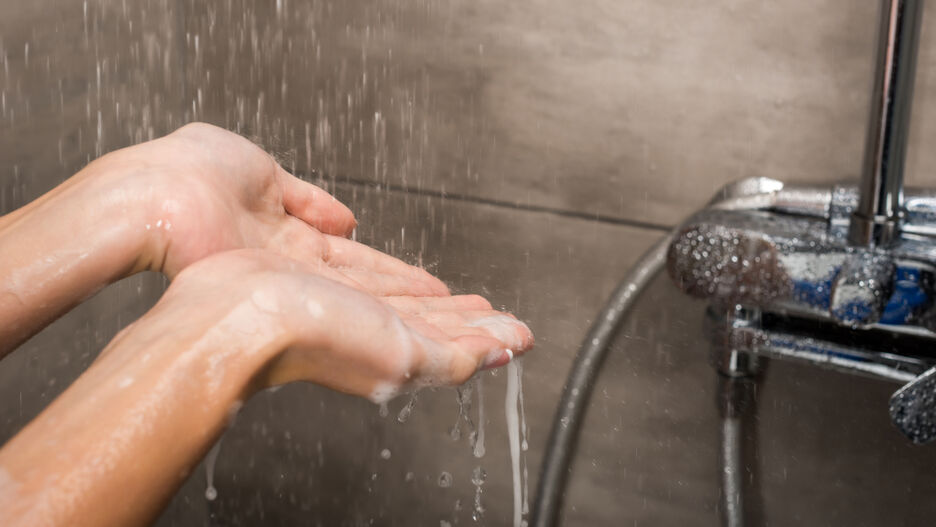 Nahaufnahme einer Dusche mit laufendem Wasser. Eine Frau hält ihre Hände unter das laufende Wasser.