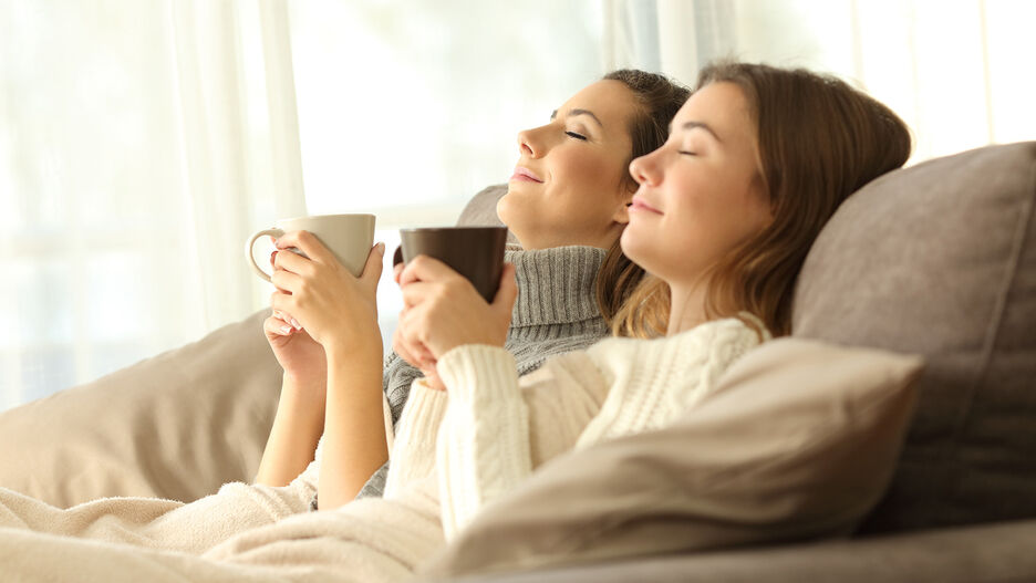 Zwei junge Frauen liegen entspannt mit einer Tasse Tee auf der Couch.